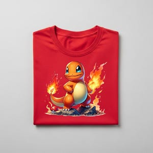 Peut inclure: T-shirt rouge pli&eacute; avec un personnage de dessin anim&eacute; Charmander avec des flammes et une Pok&eacute; Ball. Le Charmander orange a les yeux bleus et se tient sur des rochers. Les flammes sont jaunes et oranges.