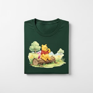 May include: Dark green t-shirt featuring a colorful illustration of Winnie the Pooh and Piglet sitting on a log in a meadow. The cartoon characters are in a sunny outdoor scene with trees and grass.