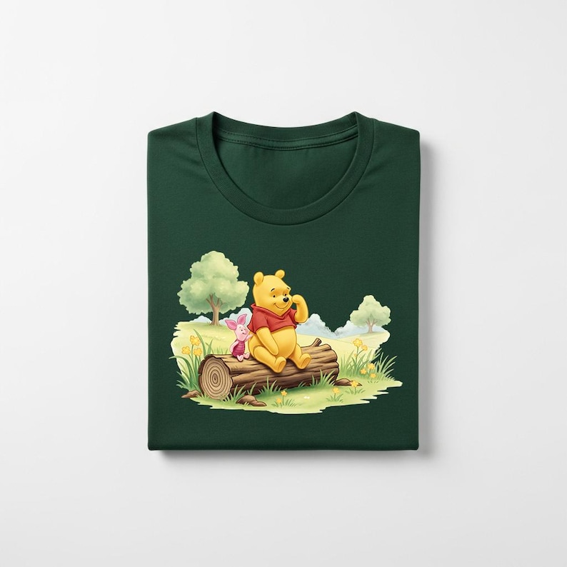 May include: Dark green t-shirt featuring a colorful illustration of Winnie the Pooh and Piglet sitting on a log in a meadow. The cartoon characters are in a sunny outdoor scene with trees and grass.
