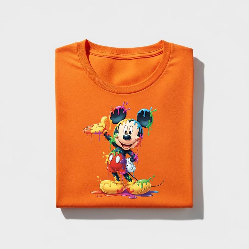May include: Orange crew neck t-shirt featuring a colorful graphic of Mickey Mouse with paint dripping from his body. The cartoon character is depicted in a playful pose, with yellow, red, blue, and green paint accents. The shirt is folded neatly.