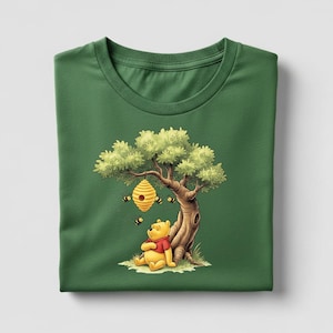 May include: Green t-shirt featuring a cartoon illustration of a yellow bear in a red shirt sitting under a tree with a beehive. Several bees are flying around the beehive. The shirt is folded neatly.