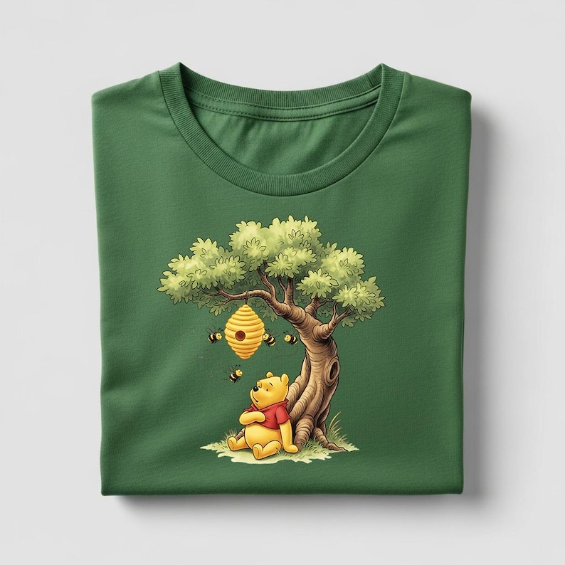 May include: Green t-shirt featuring a cartoon illustration of a yellow bear in a red shirt sitting under a tree with a beehive. Several bees are flying around the beehive. The shirt is folded neatly.