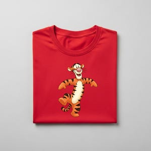 May include: A folded red t-shirt featuring a cartoon image of Tigger from Winnie the Pooh. The character is orange with black stripes, a white belly, and a pink nose. The shirt is made of a soft material.