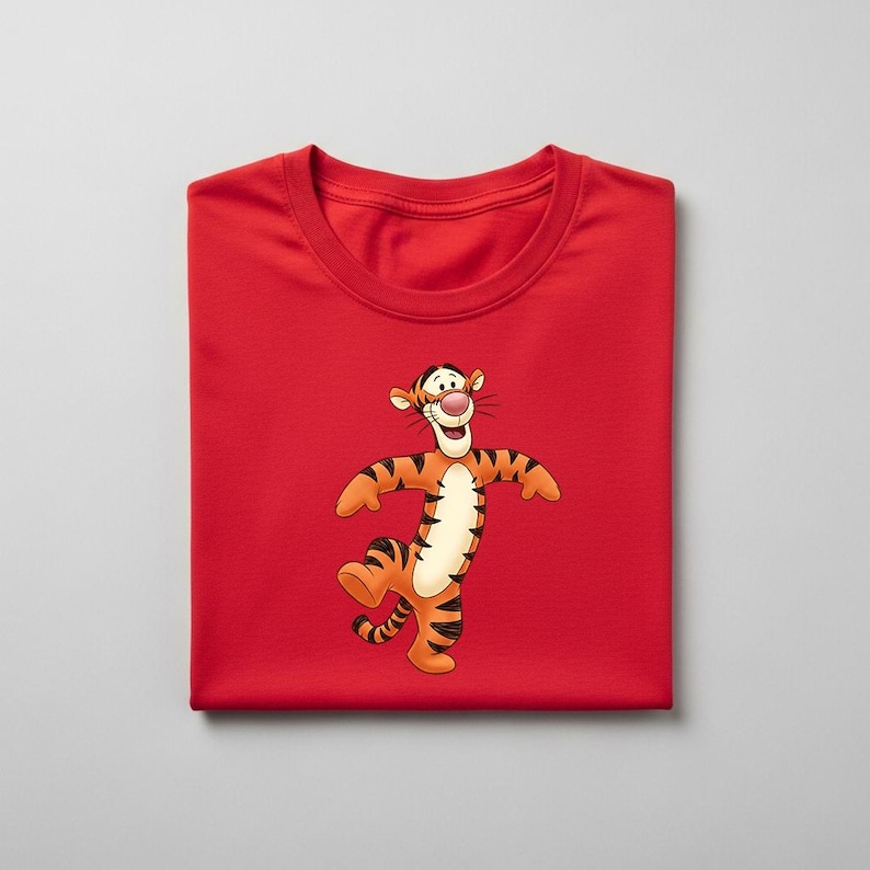 May include: A folded red t-shirt featuring a cartoon image of Tigger from Winnie the Pooh. The character is orange with black stripes, a white belly, and a pink nose. The shirt is made of a soft material.