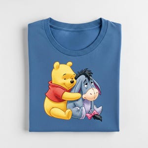 May include: A blue t-shirt featuring a graphic of Winnie the Pooh hugging Eeyore. Winnie the Pooh is yellow and wearing a red shirt. Eeyore is gray with a pink bow. The t-shirt is folded neatly.