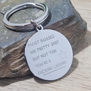 Puede incluir: Un llavero plateado con un colgante redondo. El colgante tiene el texto "MOST BOSSES ARE PRETTY SHIT BUT NOT YOU YOU'RE A FUCKING LEGEND" y una estrella. El llavero está sobre una piedra gris.