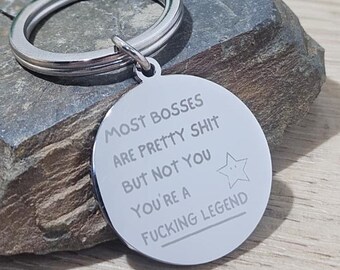 May include: A silver-colored keychain with a round pendant. The pendant has the text "MOST BOSSES ARE PRETTY SHIT BUT NOT YOU YOU'RE A FUCKING LEGEND" and a star graphic. The keychain is on a gray stone surface.