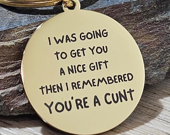 May include: A gold-colored round keychain pendant with the engraved text: "I WAS GOING TO GET YOU A NICE GIFT THEN I REMEMBERED YOU'RE A CUNT." The keychain has a gold ring.