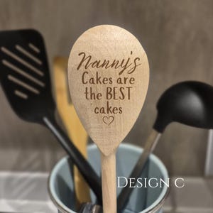 May include: A light-colored wooden spoon with the engraved text Nanny's Cakes are the BEST cakes and a small heart symbol. The spoon is positioned among other kitchen utensils in a container, with the text DESIGN C visible nearby.