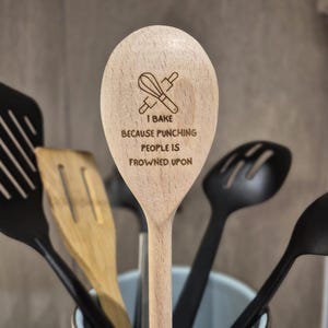 Puede incluir: Cuchara de cocina de madera con el texto "I bake because punching people is frowned upon." La cuchara tiene un dise&ntilde;o grabado con l&aacute;ser de un batidor y un rodillo. Otros utensilios de cocina negros est&aacute;n en el fondo.