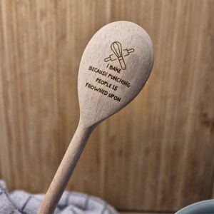 Puede incluir: Cuchara de cocina de madera clara con el texto "I BAKE BECAUSE PUNCHING PEOPLE IS FROWNED UPON" y un gr&aacute;fico de un batidor y un rodillo. Ideal para hornear y cocinar.