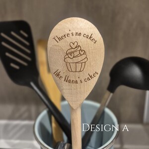 May include: Wooden spoon with a cupcake design and the text "There's no cakes like Nana's cakes." The spoon is surrounded by other kitchen utensils in a light blue container. The text "DESIGN A" is visible.