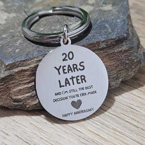 Puede incluir: Un llavero circular de metal plateado con una anilla. El colgante está grabado con el texto 20 YEARS LATER AND I'M STILL THE BEST DECISION YOU'VE EVER MADE, un pequeño icono de corazón y las palabras HAPPY ANNIVERSARY en la parte inferior.