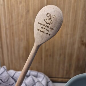 Puede incluir: Cuchara de cocina de madera clara con el texto "I BAKE BECAUSE PUNCHING PEOPLE IS FROWNED UPON" y un dibujo de un batidor y un rodillo. Perfecta para hornear y cocinar.