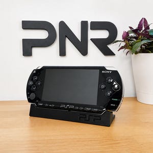 Op de afbeelding: Een zwarte Sony PSP handheld spelcomputer die in een bijpassende zwarte plastic displaystandaard staat. Het apparaat is voorzien van een breedbeeldscherm, een richtingspad, actieknoppen en het PSP-logo op de voorzijde en de standaard.