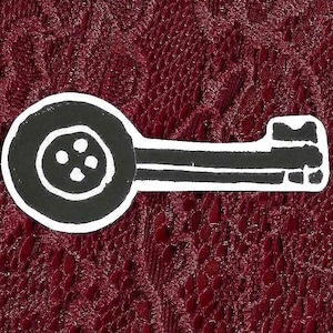 May include: A black and white graphic sticker or patch featuring a stylised key design. The head of the key is shaped like a circular four-hole button, and the shaft extends to a notched bit. The design has a hand-drawn, high-contrast aesthetic.