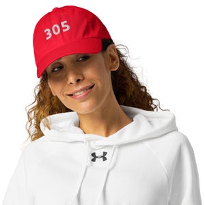 May include: A red baseball cap featuring the number "305" in white embroidery. The cap is worn by a person wearing a white hoodie.