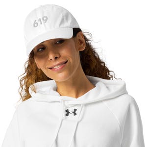 Peut inclure: Casquette de baseball blanche avec le numéro "619" brodé sur le devant. La casquette est portée par une personne portant un sweat à capuche blanc avec un logo noir.