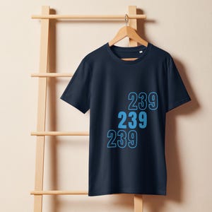Puede incluir: Camiseta azul marino con el número "239" en azul claro, repetido tres veces. La camiseta está colgada de una percha de madera. El fondo es de color beige claro, con una escalera de madera.