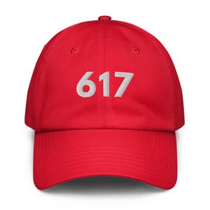 May include: A red baseball cap with the number "617" embroidered in white on the front. The cap has a curved brim and an adjustable strap at the back. The hat is made of a soft, durable fabric.
