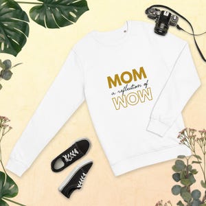 Puede incluir: Sudadera blanca de manga larga con el texto "MOM a reflection of WOW" en letras doradas. Zapatillas deportivas blancas y negras en la parte inferior izquierda. Una cámara negra en la parte superior derecha. Hojas verdes y flores rosas en el fondo.