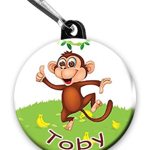 May include: A round, white zipper pull with a cartoon monkey wearing a brown shirt and holding a banana. The monkey is surrounded by green grass and yellow bananas. The text "Toby" is written in brown letters at the bottom of the pull.