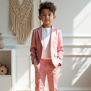 Puede incluir: Un traje de color rosa empolvado con chaqueta cruzada y pantalón a juego, usado sobre una camiseta blanca lisa. La chaqueta presenta solapas de muesca, dos botones dorados en la parte delantera y bolsillos con solapa. Se combina con zapatillas blancas.