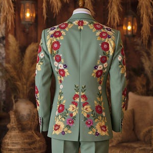 May include: A sage green suit jacket and pants set with intricate floral embroidery. The embroidery features red, yellow, white, and purple flowers with green leaves. The suit is tailored and appears to be made of a smooth fabric.