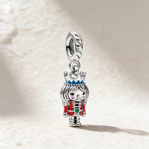 May include: A silver charm featuring a Nutcracker soldier design. The charm has a red jacket, blue crown, and a green heart. The charm is suspended from a silver loop.