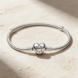 Puede incluir: Un brazalete plateado con un dije en forma de corazón con la inscripción "Pandora". El brazalete tiene un diseño texturizado y flexible, presentado sobre una superficie clara. El brazalete mide aproximadamente 18 cm de largo.