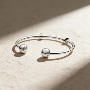 May include: A silver open bangle bracelet with two spherical charms and two small cylindrical accents. The charms have engraved text. The bracelet is displayed on a light-coloured surface, with shadows.
