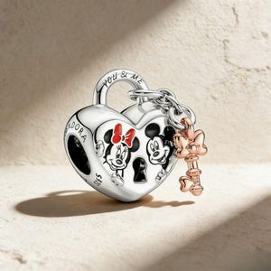 May include: A silver heart-shaped charm with Mickey and Minnie Mouse illustrations. The charm has a key and the words "YOU & ME" engraved on it. The key is rose gold with a bow design.