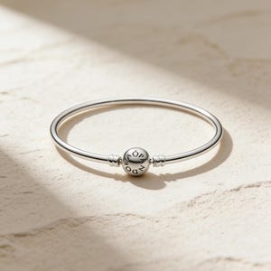 May include: A silver bangle bracelet with a central round charm. The charm is engraved with the word "Pandora". The bracelet has a simple, elegant design, suitable for everyday wear. The bracelet's diameter is approximately 6 cm.