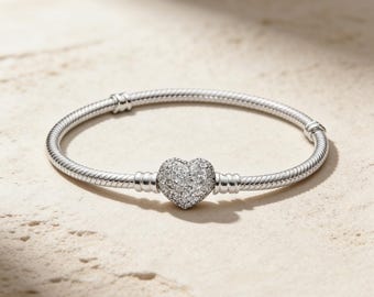 May include: A silver bracelet with a heart-shaped charm. The charm is covered in small, sparkling stones. The bracelet is on a light-colored surface.