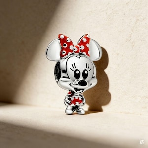 May include: A silver Minnie Mouse charm with a red bow and dress, both decorated with white polka dots. The charm features a smiling face and is a collectible item.