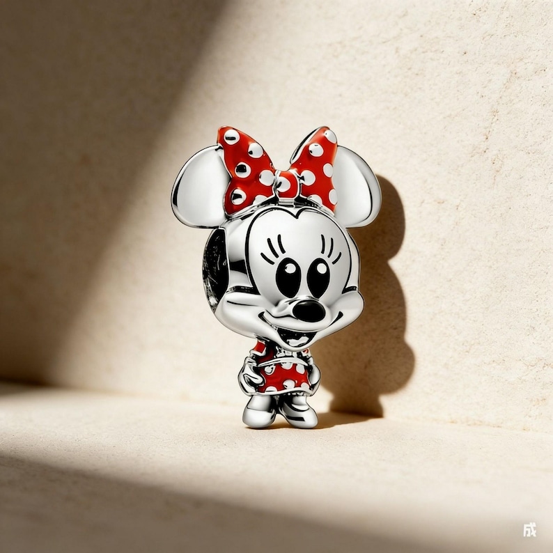 May include: A silver Minnie Mouse charm with a red bow and dress, both decorated with white polka dots. The charm features a smiling face and is a collectible item.
