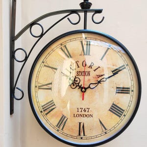 Peut inclure: Horloge murale double face avec un cadre et un support en métal noir. Le cadran de l'horloge présente des chiffres romains et le texte "Victoria Station 1747 London". Les aiguilles sont noires avec une trotteuse rouge.