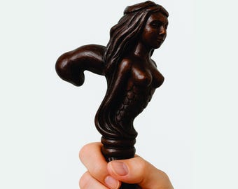 Puede incluir: Bastón de madera marrón oscuro con una figura de sirena tallada en la parte superior. La sirena tiene cabello largo y fluido y una cola de pez detallada. El bastón se sostiene en una mano sobre un fondo blanco.