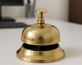 May include: A polished brass service bell with a smooth, rounded dome top and a central push-button plunger. The bell features a wide, flared circular base and a metallic gold finish that reflects light across its curved, reflective surface.