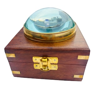 Puede incluir: Una lupa con una lente clara y abovedada, engarzada en un anillo de latón, colocada sobre una caja de madera oscura. La caja tiene esquinas de latón y un cierre de latón. La lupa es de estilo vintage.