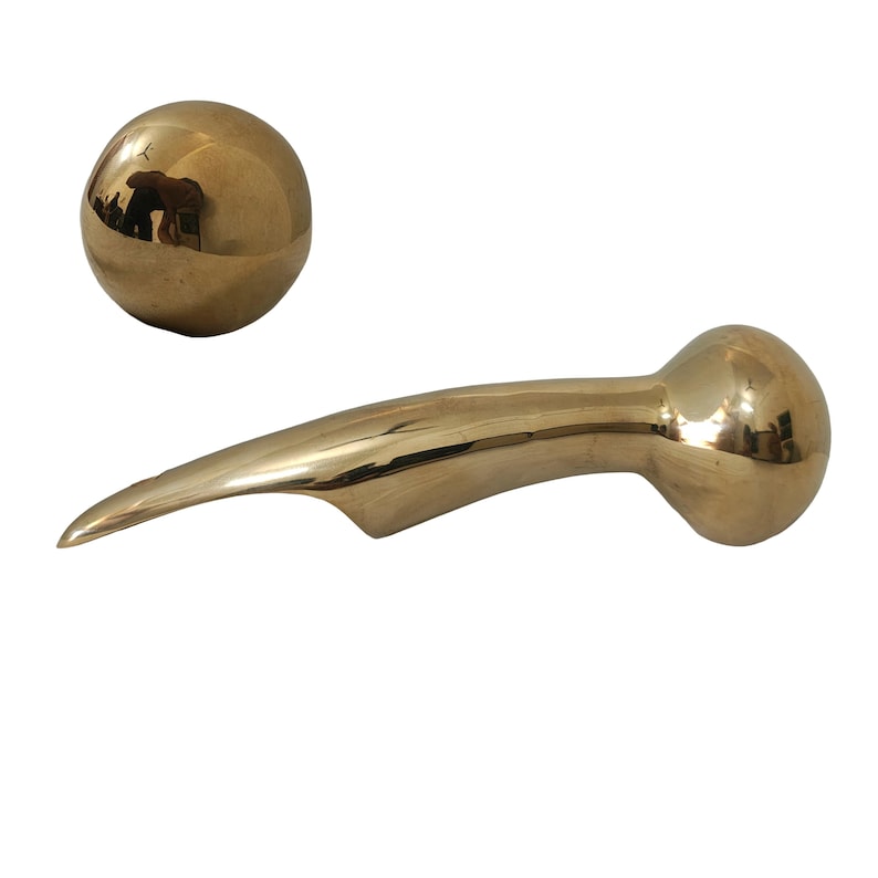 Puede incluir: Un abridor de botellas de metal dorado con un dise&ntilde;o curvo y puntiagudo, acompa&ntilde;ado de una esfera dorada. El abridor tiene un extremo bulboso y un cuerpo elegante y alargado, lo que sugiere una est&eacute;tica moderna.