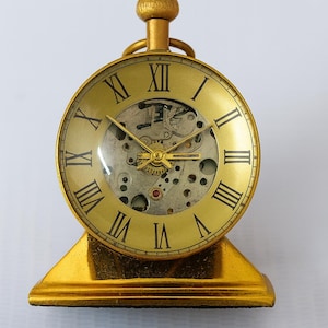 Puede incluir: Un reloj de escritorio dorado de estilo vintage, con números romanos en la esfera. El reloj tiene una ventana transparente que revela el mecanismo interno y se apoya en una base dorada. El reloj mide aproximadamente 10 cm de alto.