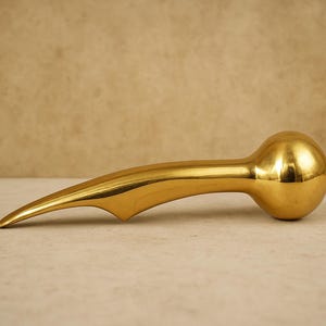Puede incluir: Un tirador de puerta decorativo dorado pulido con un extremo redondeado y bulboso y una punta curva y puntiaguda. El tirador tiene una superficie lisa y reflectante, sobre un fondo neutro.