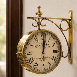 May include: A double-sided wall-mounted clock with a polished brass finish. The off-white clock face features black Roman numerals and the text Victoria Station Railway Clock London. It is supported by a decorative brass bracket with scrollwork.