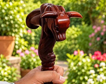 Puede incluir: Un mango de bastón de madera de color marrón oscuro tallado con la forma de una cabeza de toro con cuernos prominentes. La madera presenta un acabado pulido y detalles intrincados en la cara del toro y en la sección del cuello del mango.