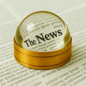 Puede incluir: Un pisapapeles de cristal en forma de cúpula con una base de metal dorado sobre una página impresa. El cristal transparente amplía el texto inferior, mostrando las palabras The News en una fuente negra con remates sobre texto lorem ipsum.
