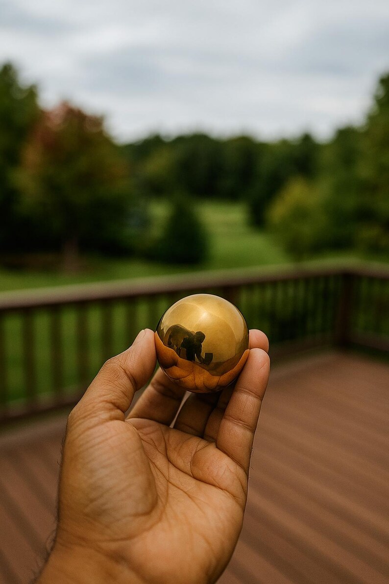 Puede incluir: Una esfera dorada y brillante sostenida en una mano. La esfera refleja el entorno, incluyendo &aacute;rboles y una persona. El fondo presenta una terraza de madera y un paisaje natural verde.