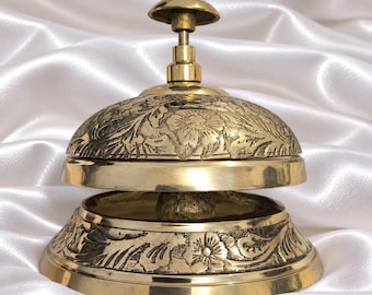 May include: A polished brass desk bell featuring an intricate floral engraving across the dome and base. The bell has a smooth, rounded top plunger and a wide, flared bottom edge, showcasing a metallic gold finish with detailed etched patterns.