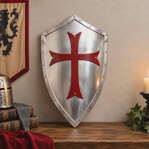 May include: A polished silver-toned metal heater shield featuring a prominent red cross emblem in the center. The shield has a pointed base and is bordered by a series of small, evenly spaced silver rivets along the entire outer edge.