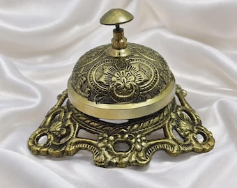 Puede incluir: Un timbre de servicio de metal color latón con un patrón floral en relieve en la cúpula y una base ancha y decorativa con detalles recortados. El timbre tiene un pequeño botón pulsador redondeado en la parte superior y acabado pulido.